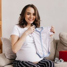 A pregnant woman with a joyful expression holding her baby bump, surrounded by an assortment of baby products such as clothing, toys, and a crib. The image represents the act of purchasing items for an unborn baby, symbolizing the excitement and preparation for their arrival.
