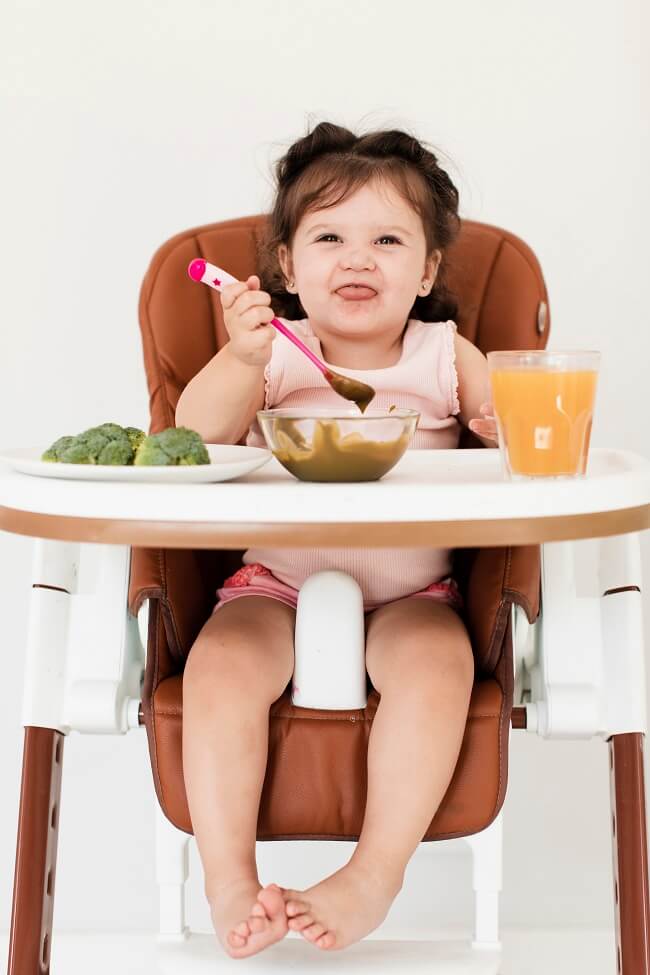 Picture of a baby sitting in a high chair, being fed mashed fruits or vegetables as they begin the journey of introducing solid foods.