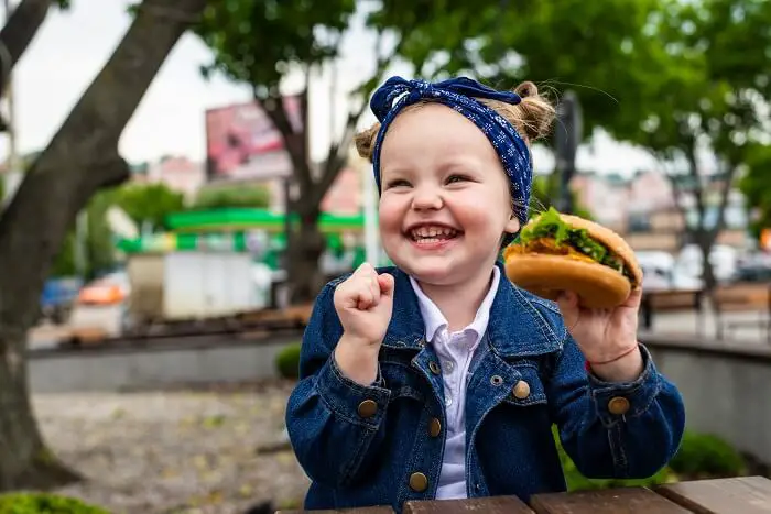 A smiling toddler enjoys a grilled cheese sandwich at a picnic table with various shaped sandwiches around.